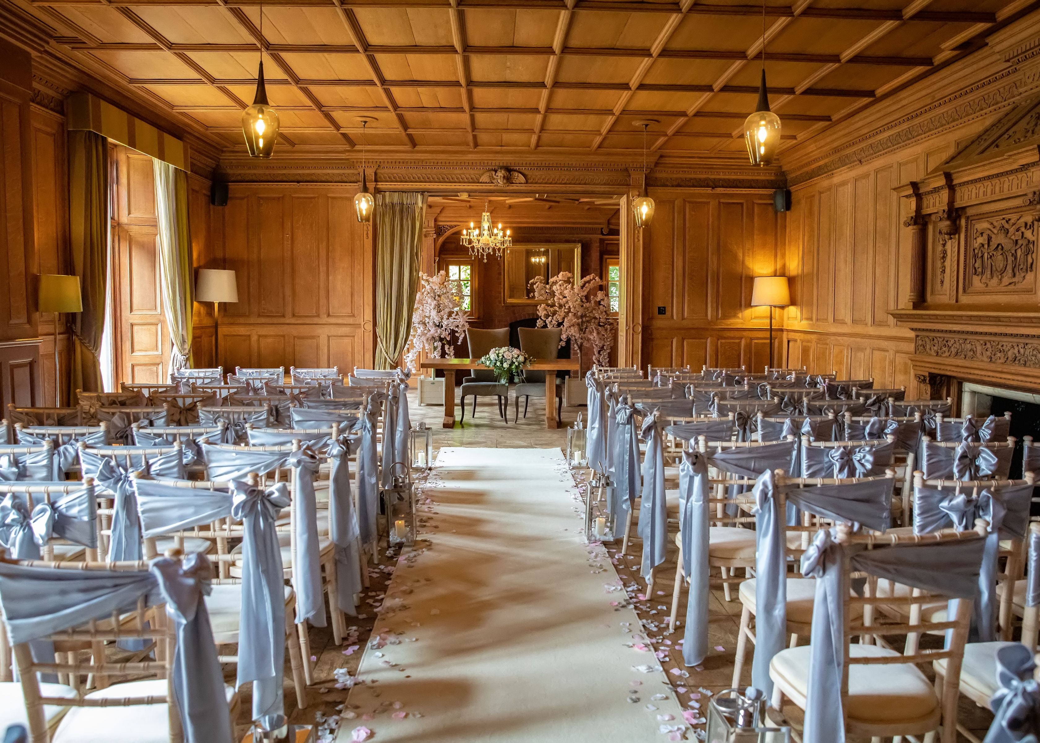 Elegant indoor wedding ceremony setup with rows of chairs draped in light gray fabric, wooden paneled walls, flower petals lining the aisle, and floral arrangements at the altar.
