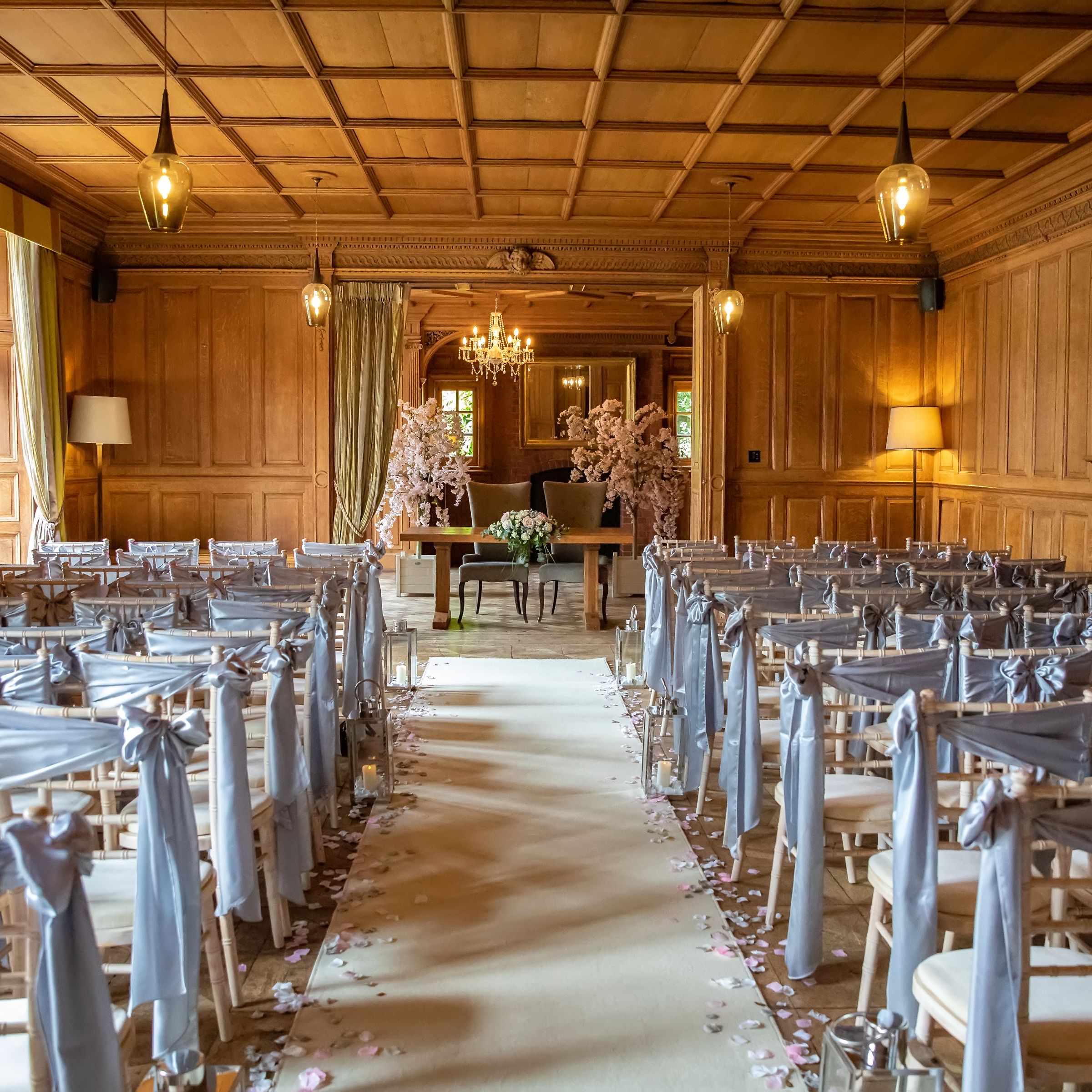 Elegant indoor wedding ceremony setup with rows of chairs draped in light gray fabric, wooden paneled walls, flower petals lining the aisle, and floral arrangements at the altar.