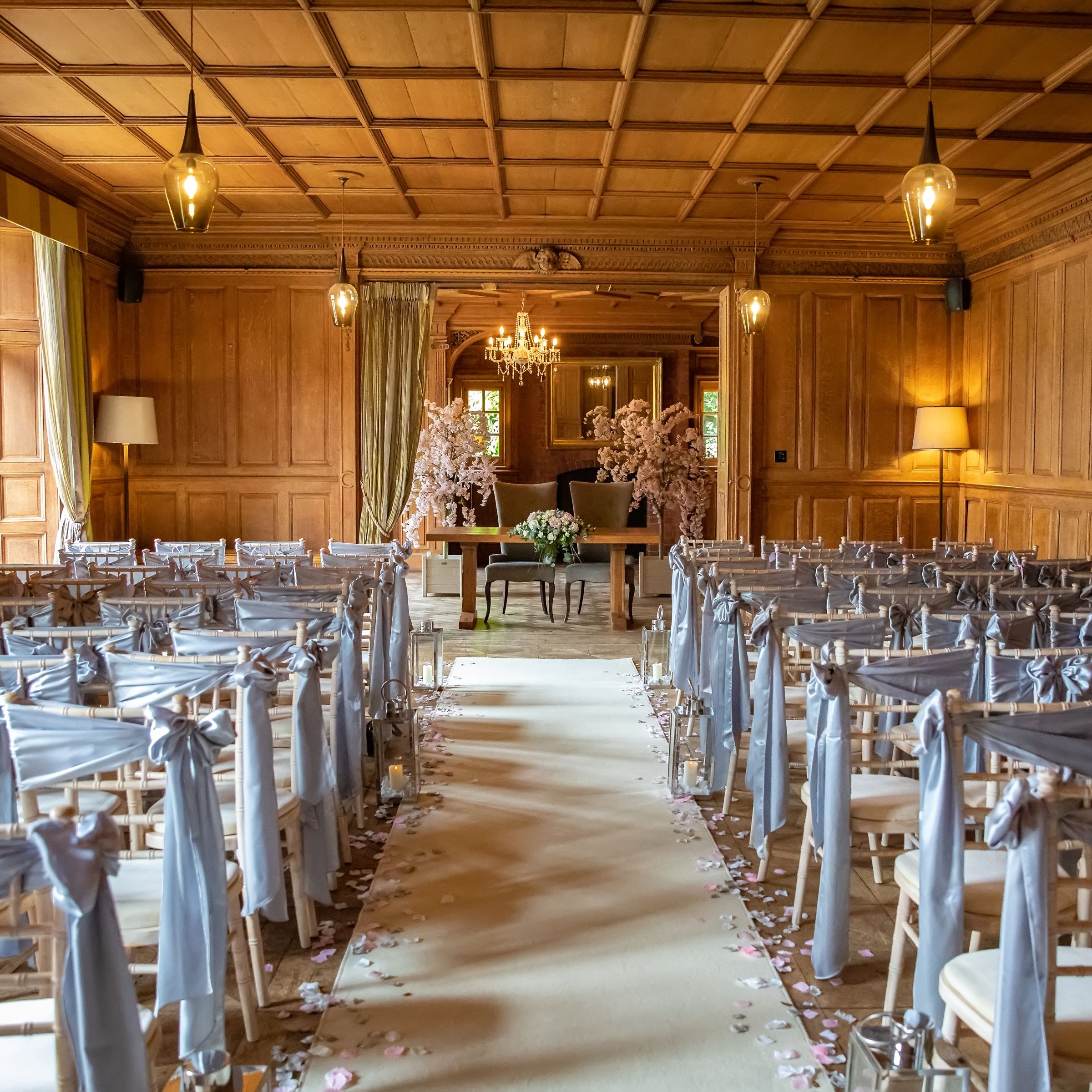 Elegant indoor wedding ceremony setup with rows of chairs draped in light gray fabric, wooden paneled walls, flower petals lining the aisle, and floral arrangements at the altar.