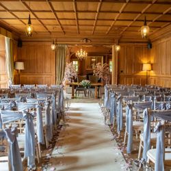 Elegant indoor wedding ceremony setup with rows of chairs draped in light gray fabric, wooden paneled walls, flower petals lining the aisle, and floral arrangements at the altar.