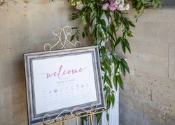 Wedding welcome sign on a stand with floral arrangement against a stone wall.