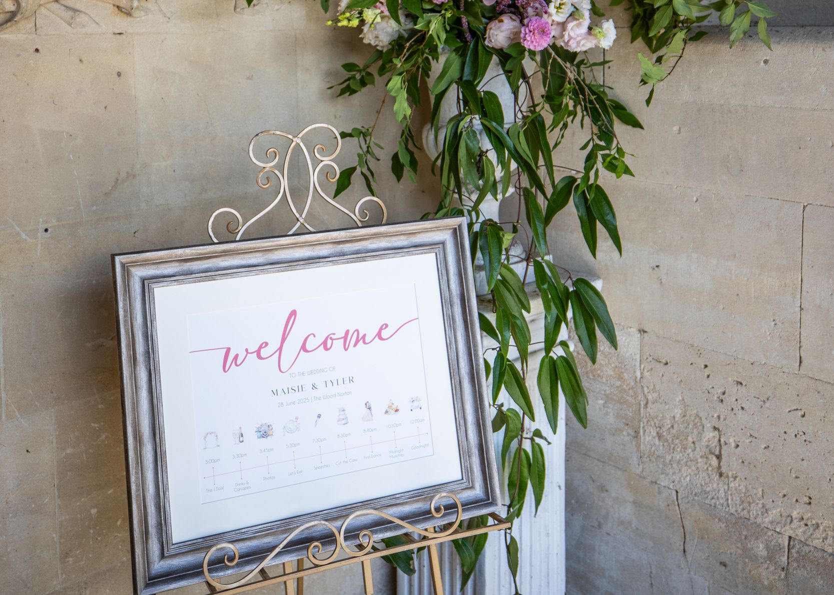 Wedding welcome sign on a stand with floral arrangement against a stone wall.