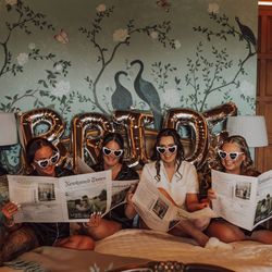 Four women wearing matching sleepwear and heart-shaped sunglasses sitting on a bed with BRIDE letter balloons behind them, reading newspapers.
