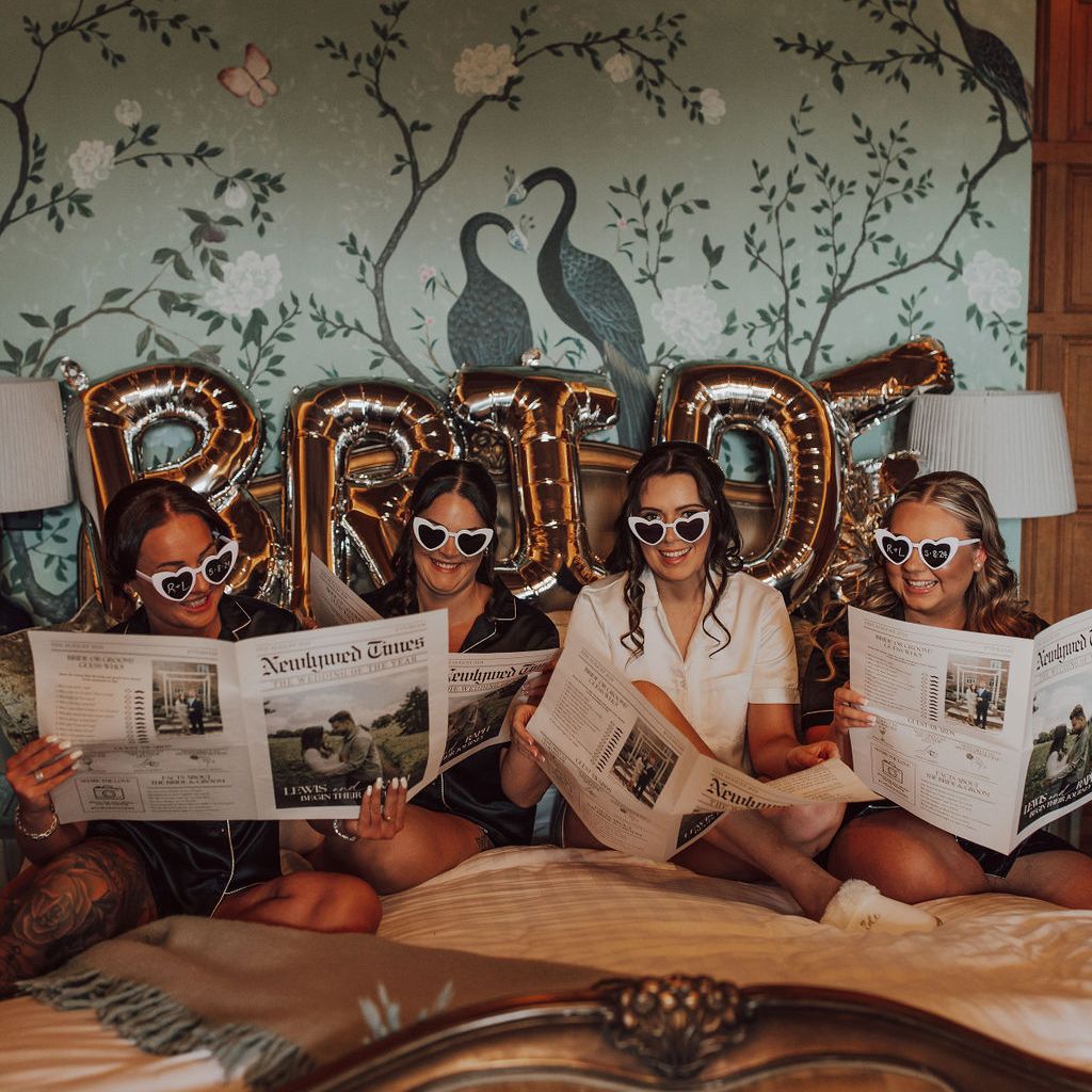 Four women wearing matching sleepwear and heart-shaped sunglasses sitting on a bed with BRIDE letter balloons behind them, reading newspapers.