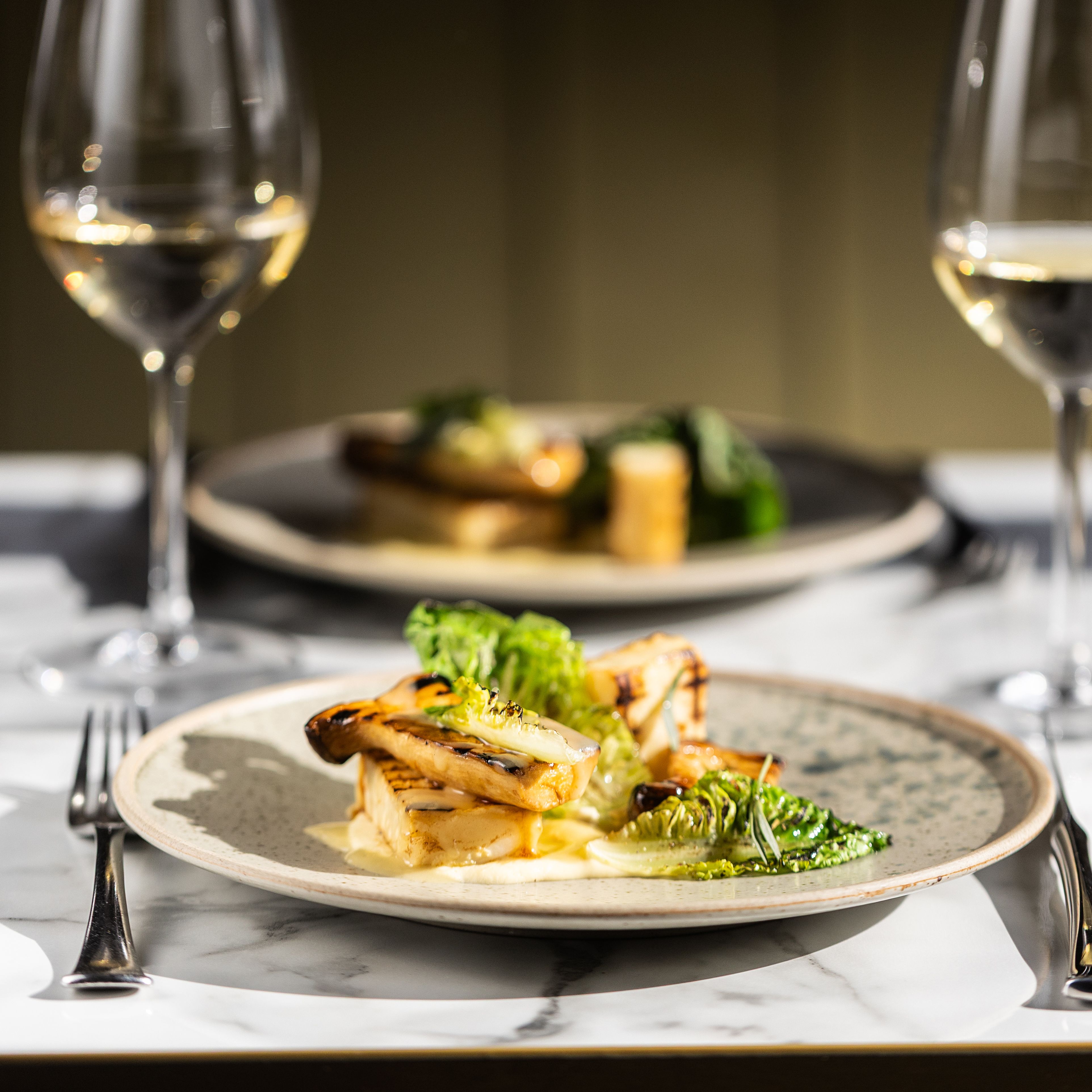 A gourmet meal plated with greens and grilled vegetables, served with white wine on a marble table.
