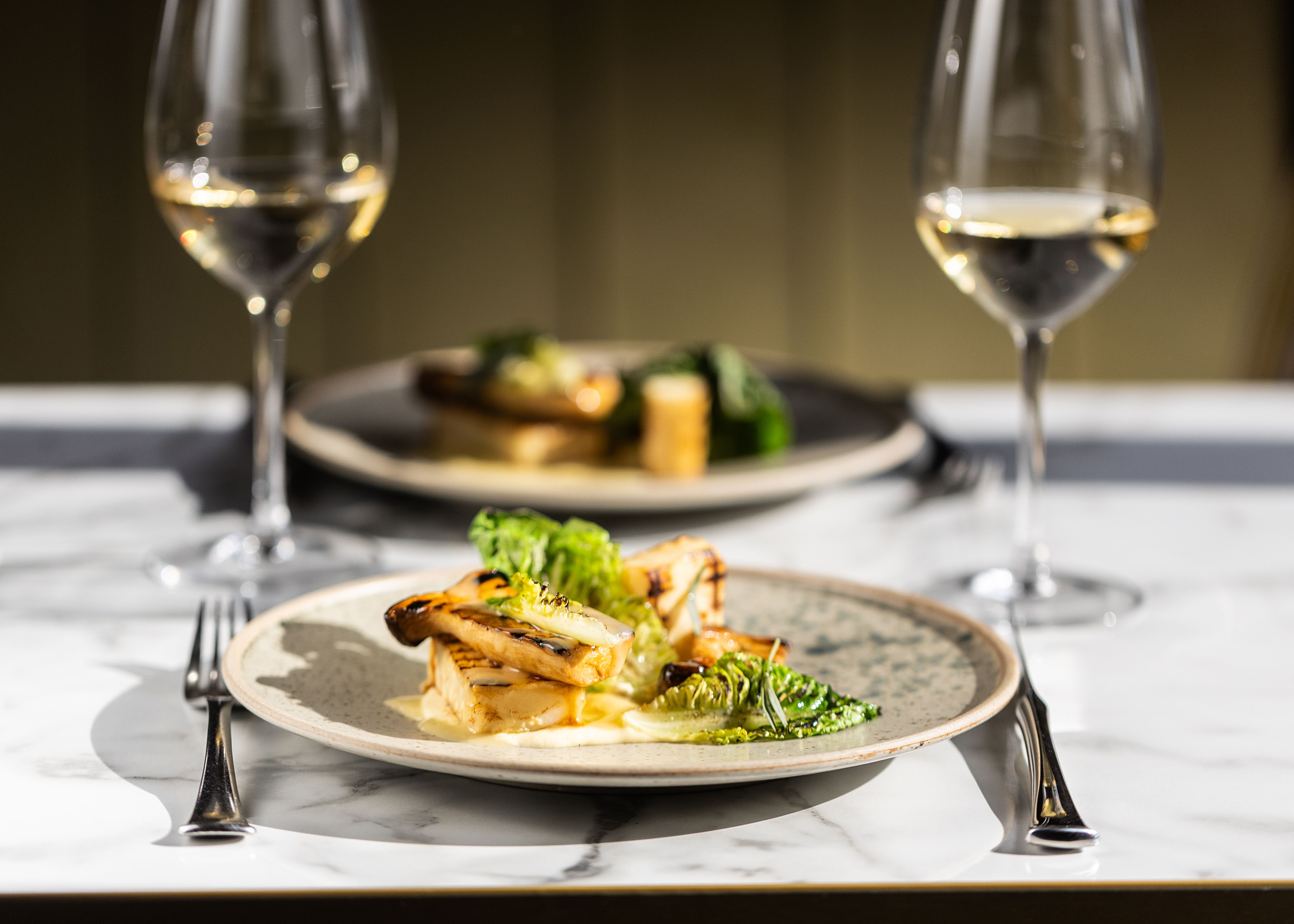A gourmet meal plated with greens and grilled vegetables, served with white wine on a marble table.