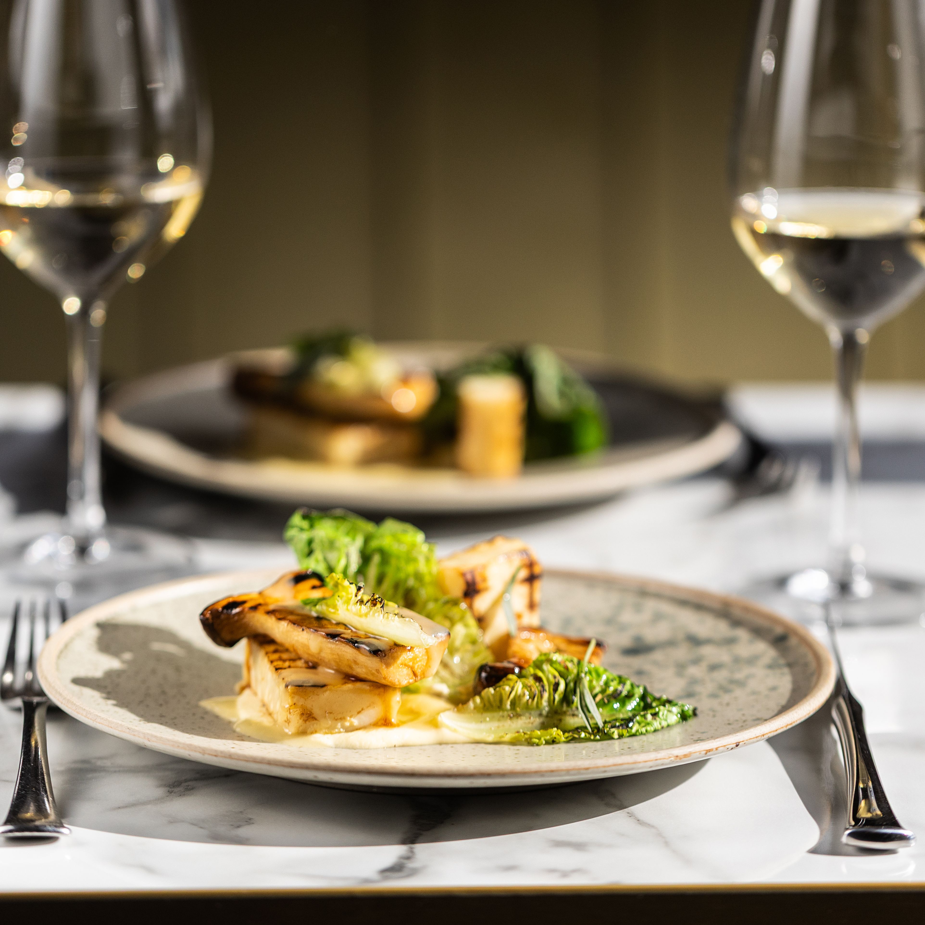 A gourmet meal plated with greens and grilled vegetables, served with white wine on a marble table.