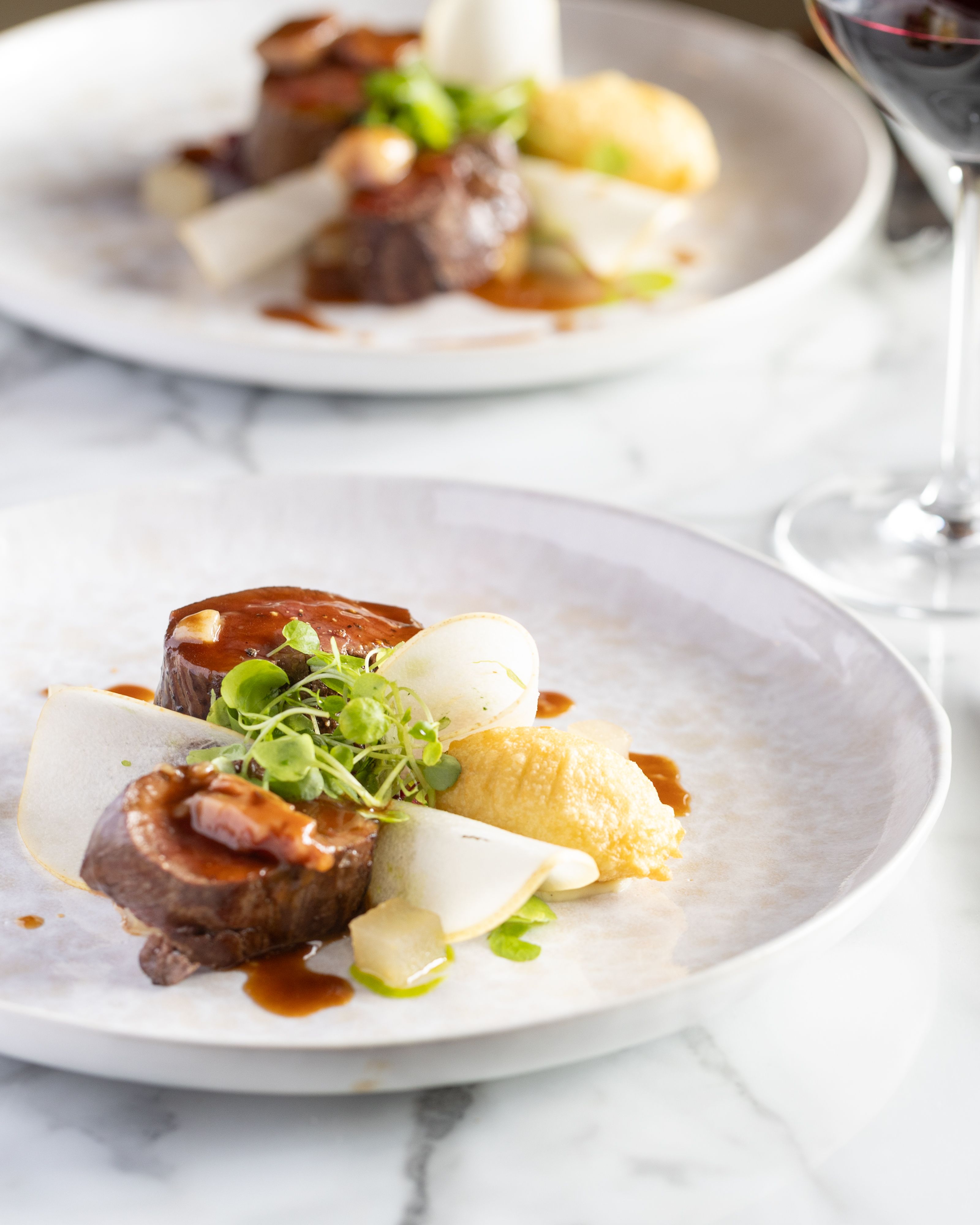 Fine dining plate featuring two pieces of meat with sauce, puree, thinly sliced vegetables, and microgreens, accompanied by a glass of red wine.
