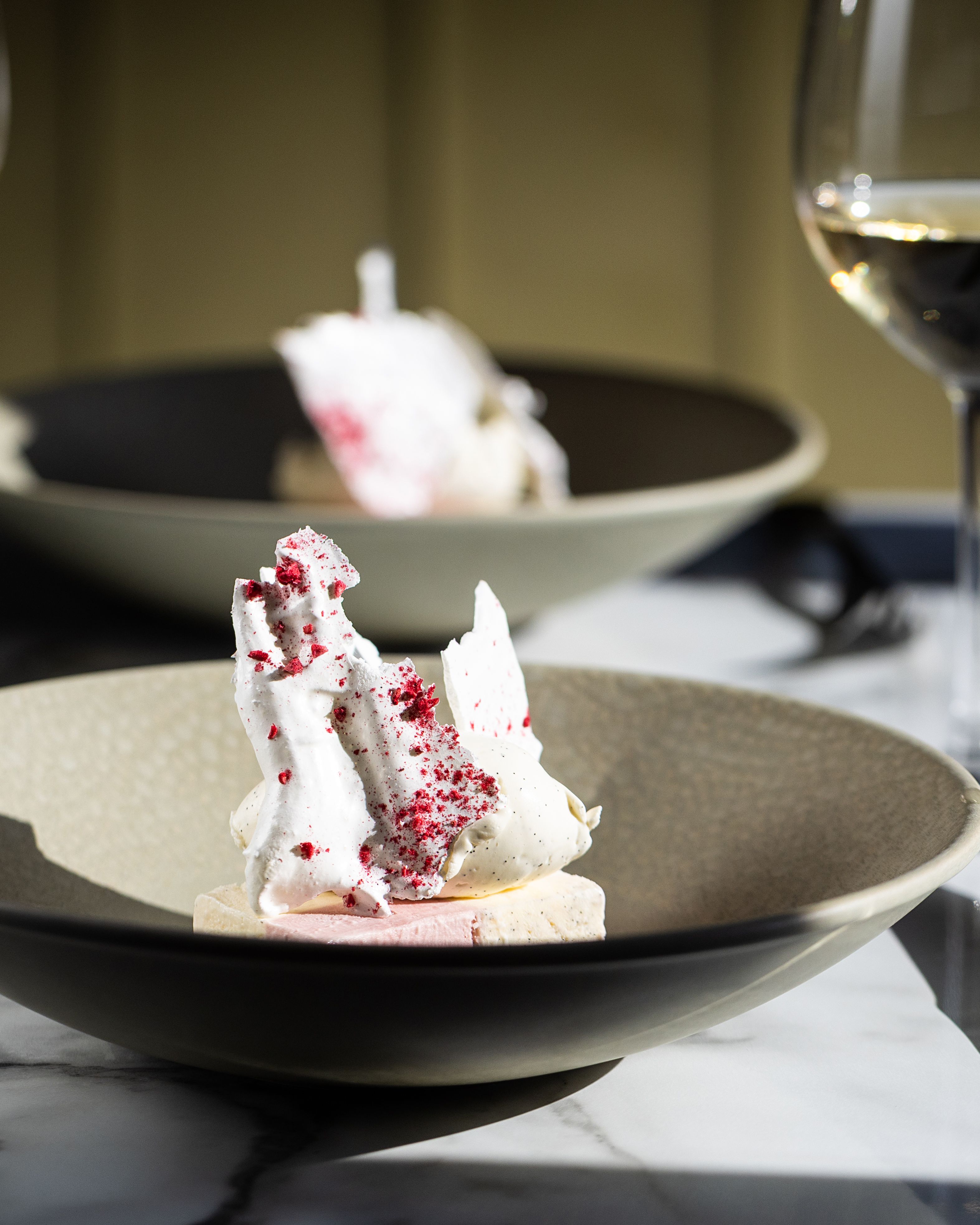 Elegant dessert with meringue and fruit pieces served in a bowl with two glasses of white wine on a marble table.