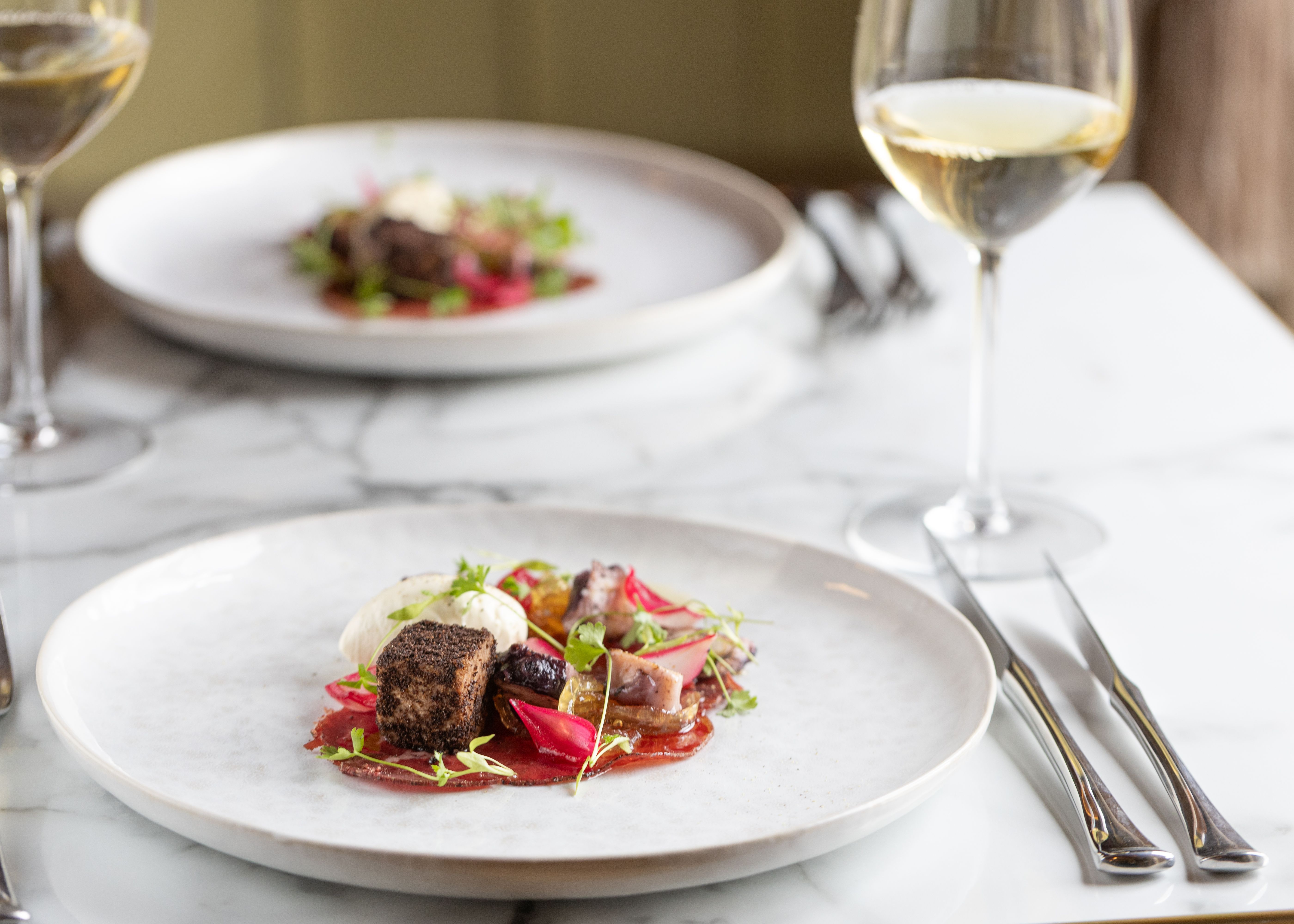 A gourmet plated dish with various colorful ingredients beside a glass of white wine on a marble table.