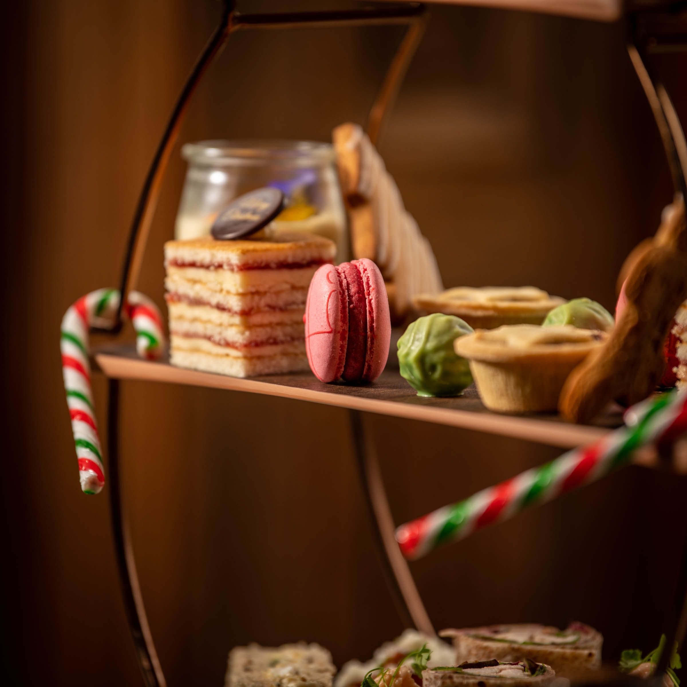 A festive afternoon tea stand with assorted desserts including a pink macaron, layered cake, mince pies, and candy canes.