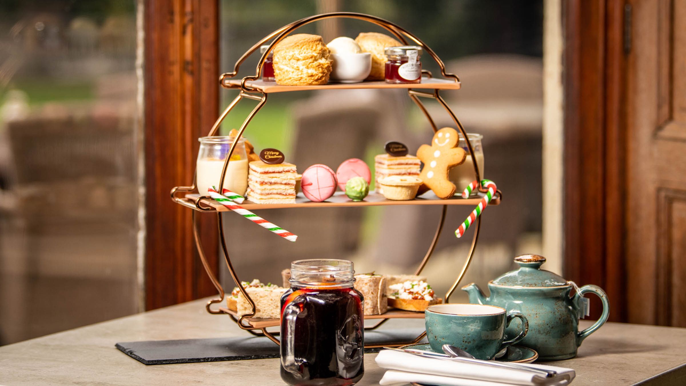 Festive afternoon tea set with teapot, cup, and a three-tiered stand of assorted pastries, sandwiches, and desserts.