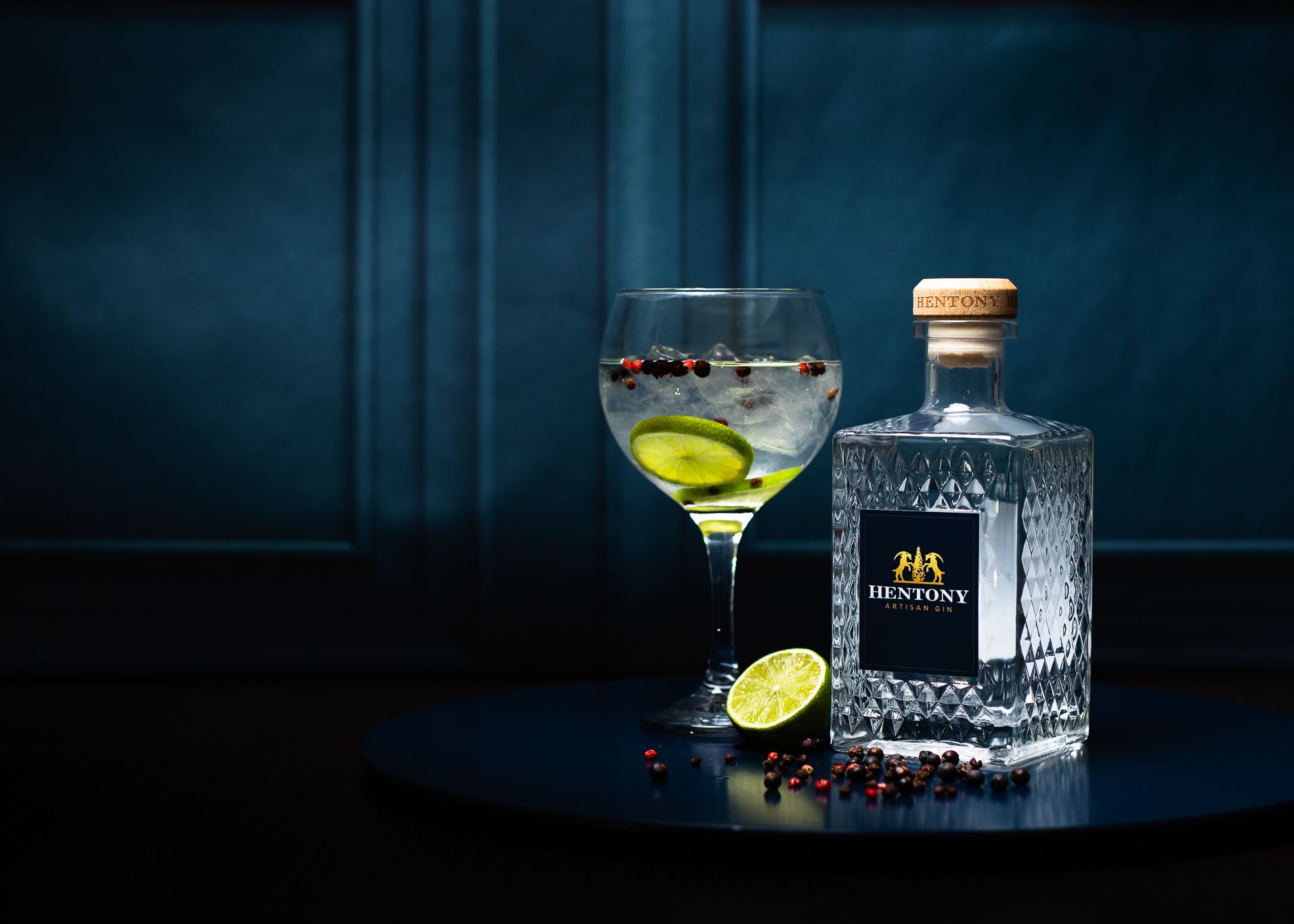 A bottle of Hentony Artisan Gin next to a glass of gin and tonic with lime and botanicals on a dark round surface.
