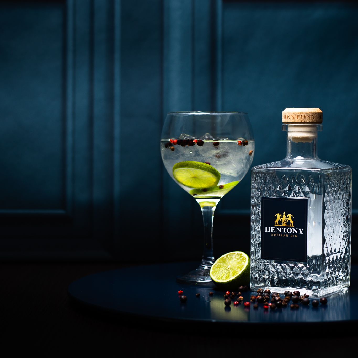 A bottle of Hentony Artisan Gin next to a glass of gin and tonic with lime and botanicals on a dark round surface.