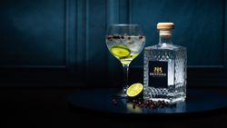 A bottle of Hentony Artisan Gin next to a glass of gin and tonic with lime and botanicals on a dark round surface.