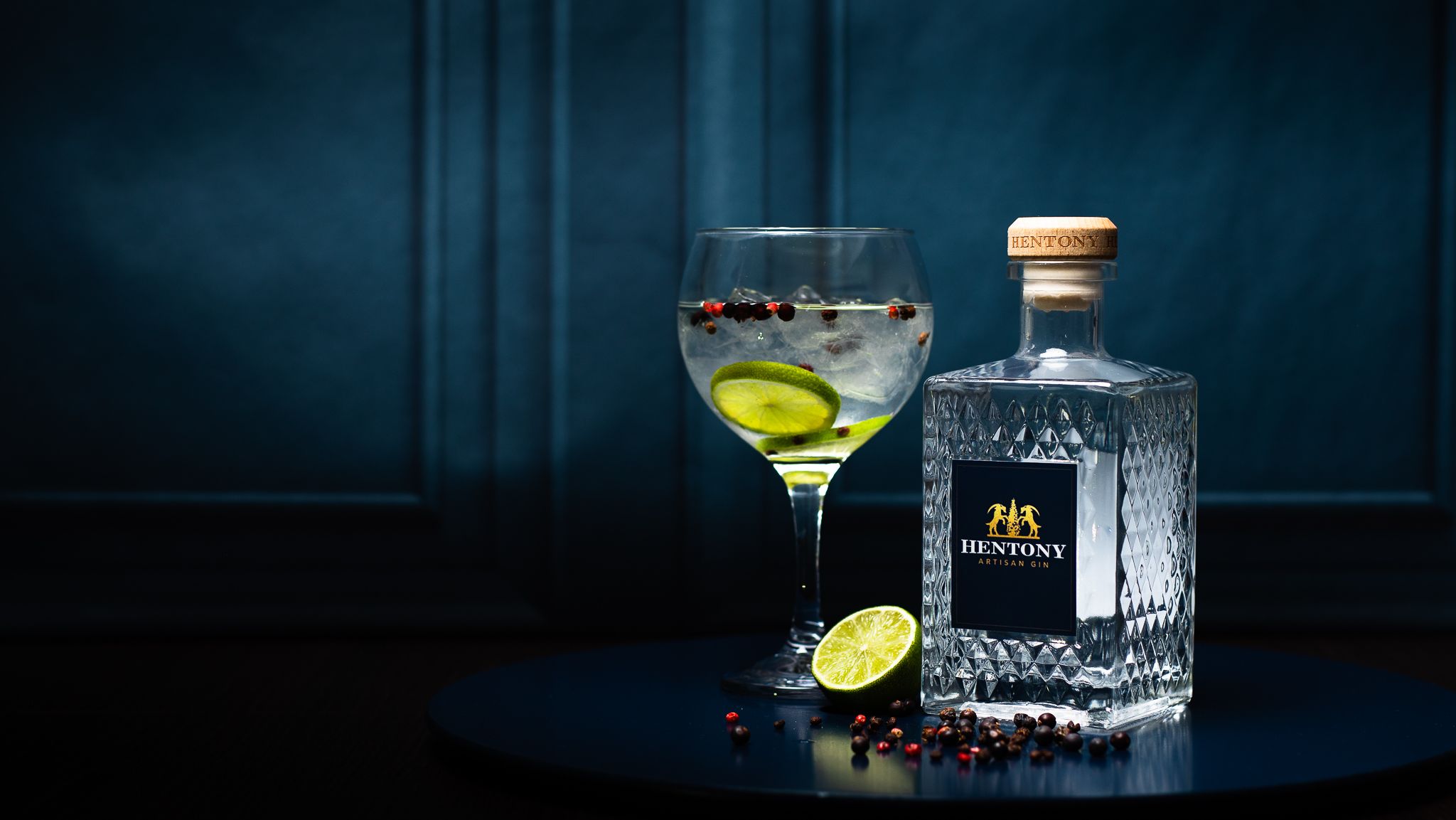 A bottle of Hentony Artisan Gin next to a glass of gin and tonic with lime and botanicals on a dark round surface.