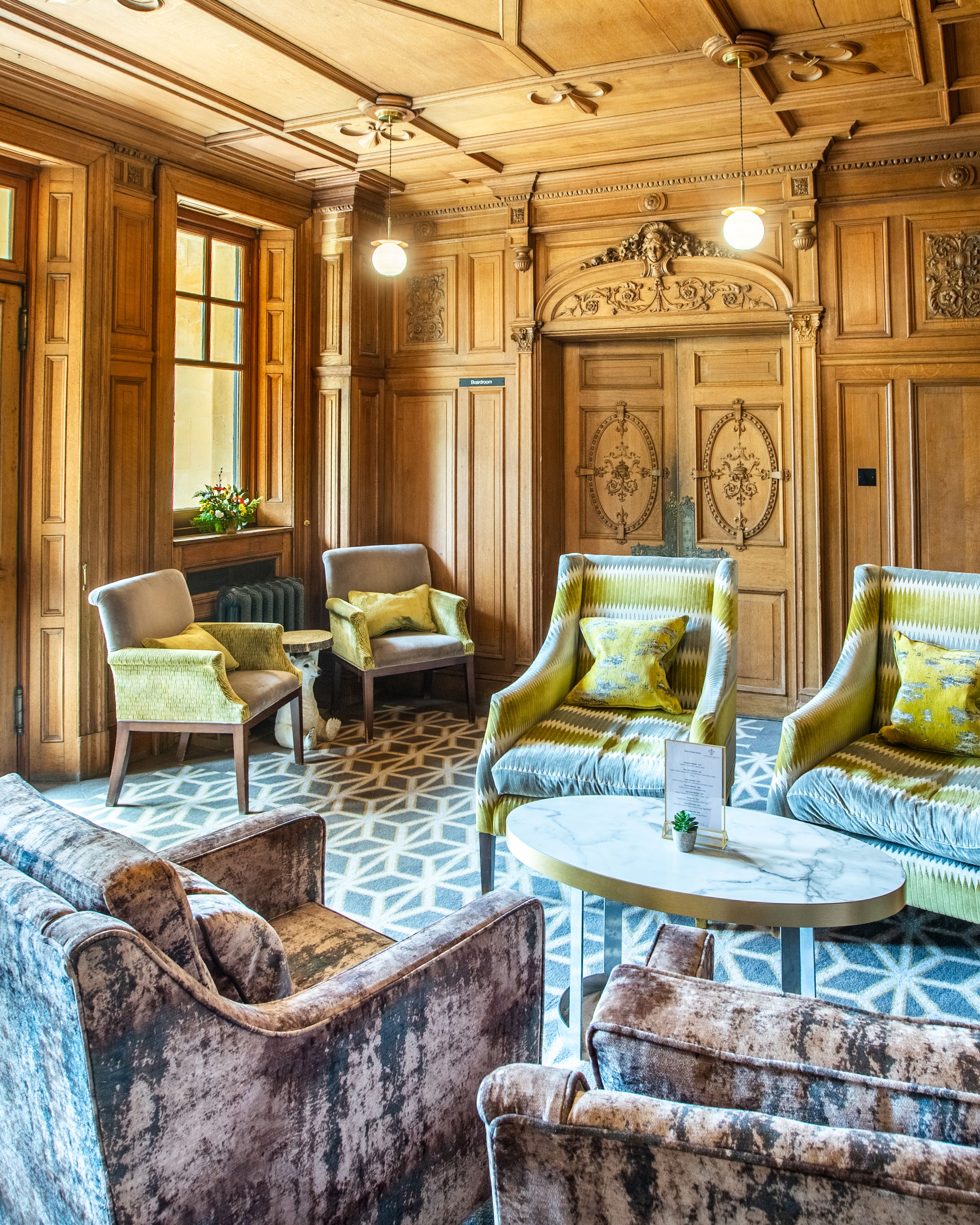 Elegant wood-paneled sitting room with velvet armchairs, ornate ceiling, and geometric carpet.