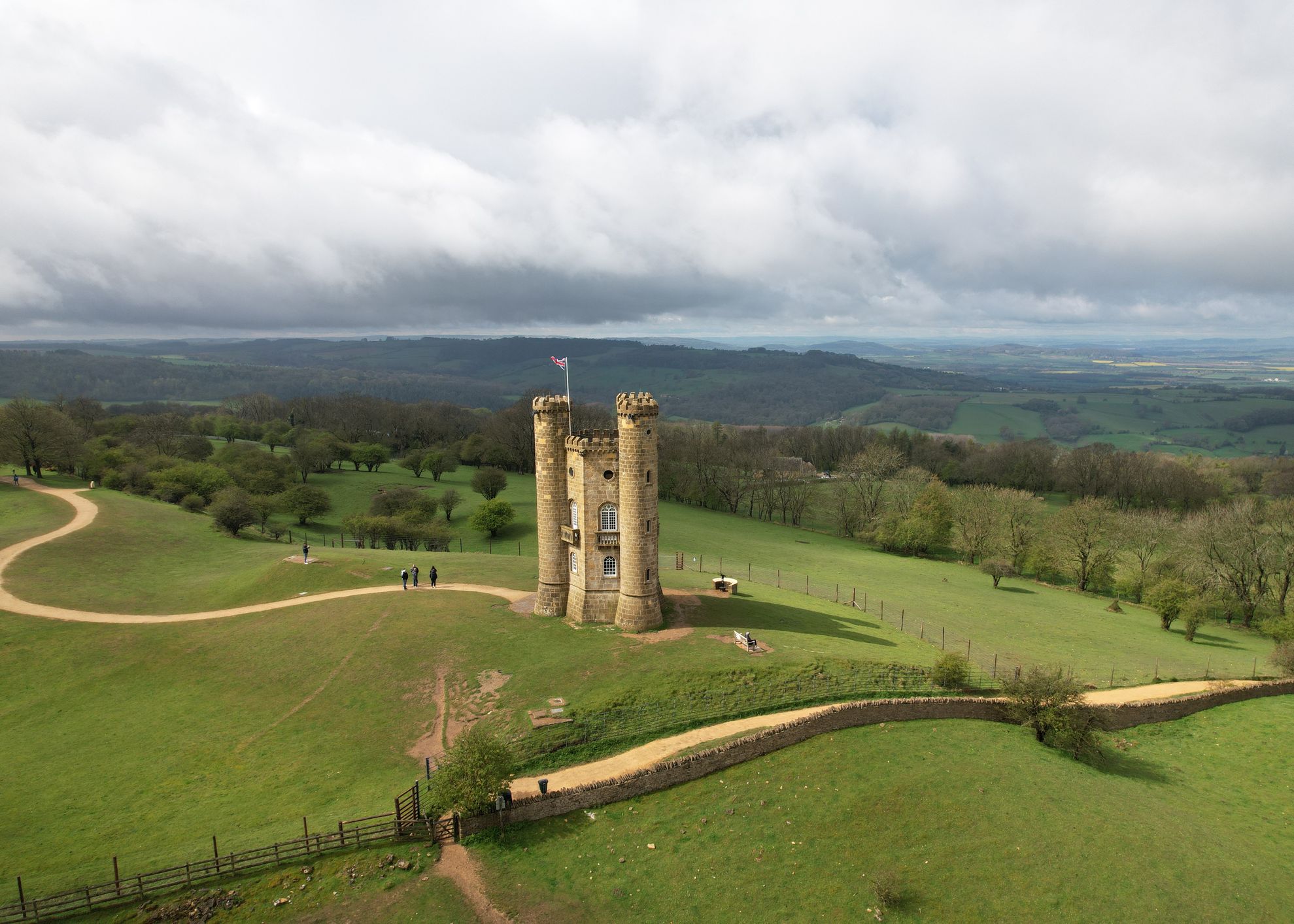 Aerial view of a small stone tower with three circular turrets on a hill surrounded by green fields and trees.