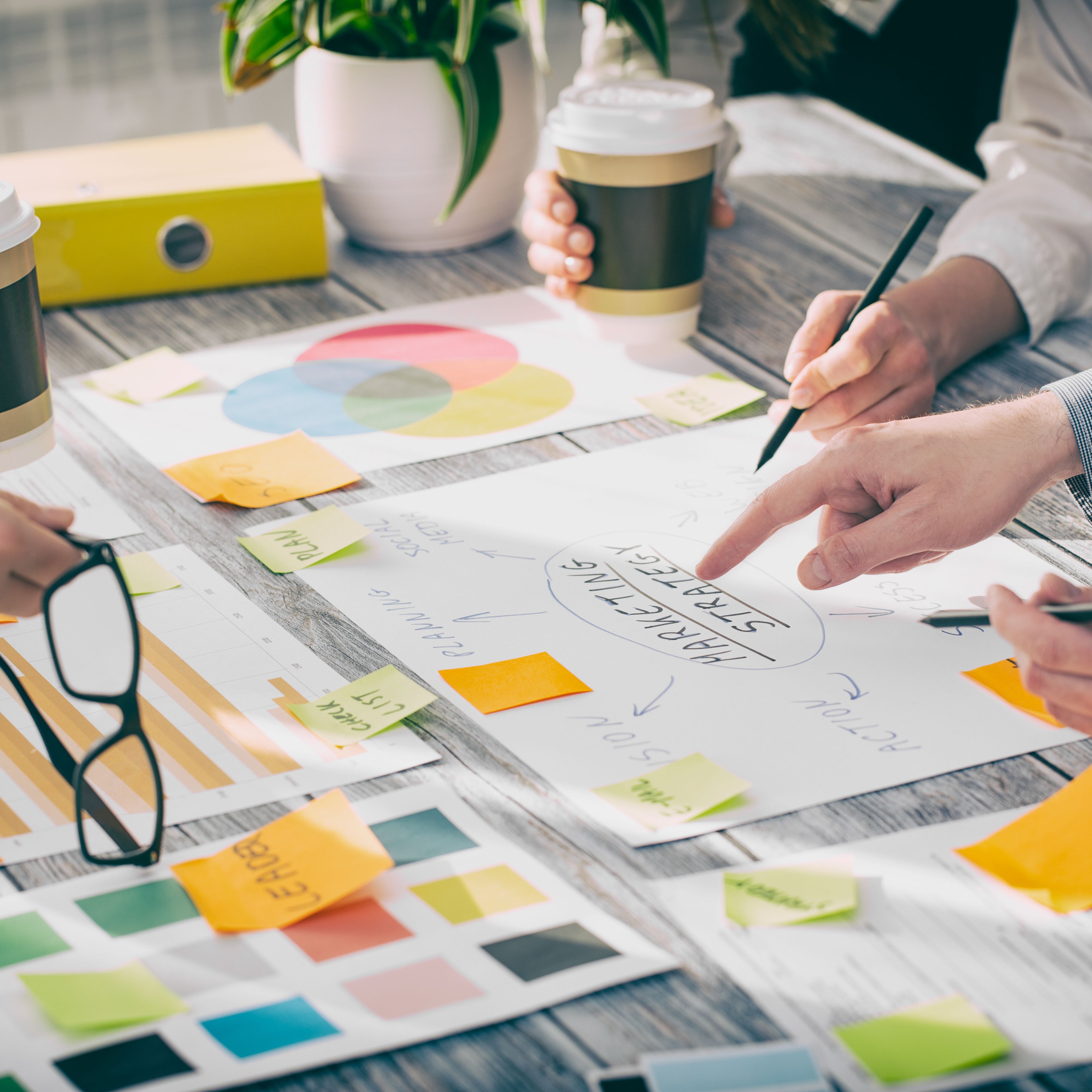 Colleagues collaborating at a desk with charts, sticky notes and takeaway coffee cups, discussing marketing strategy