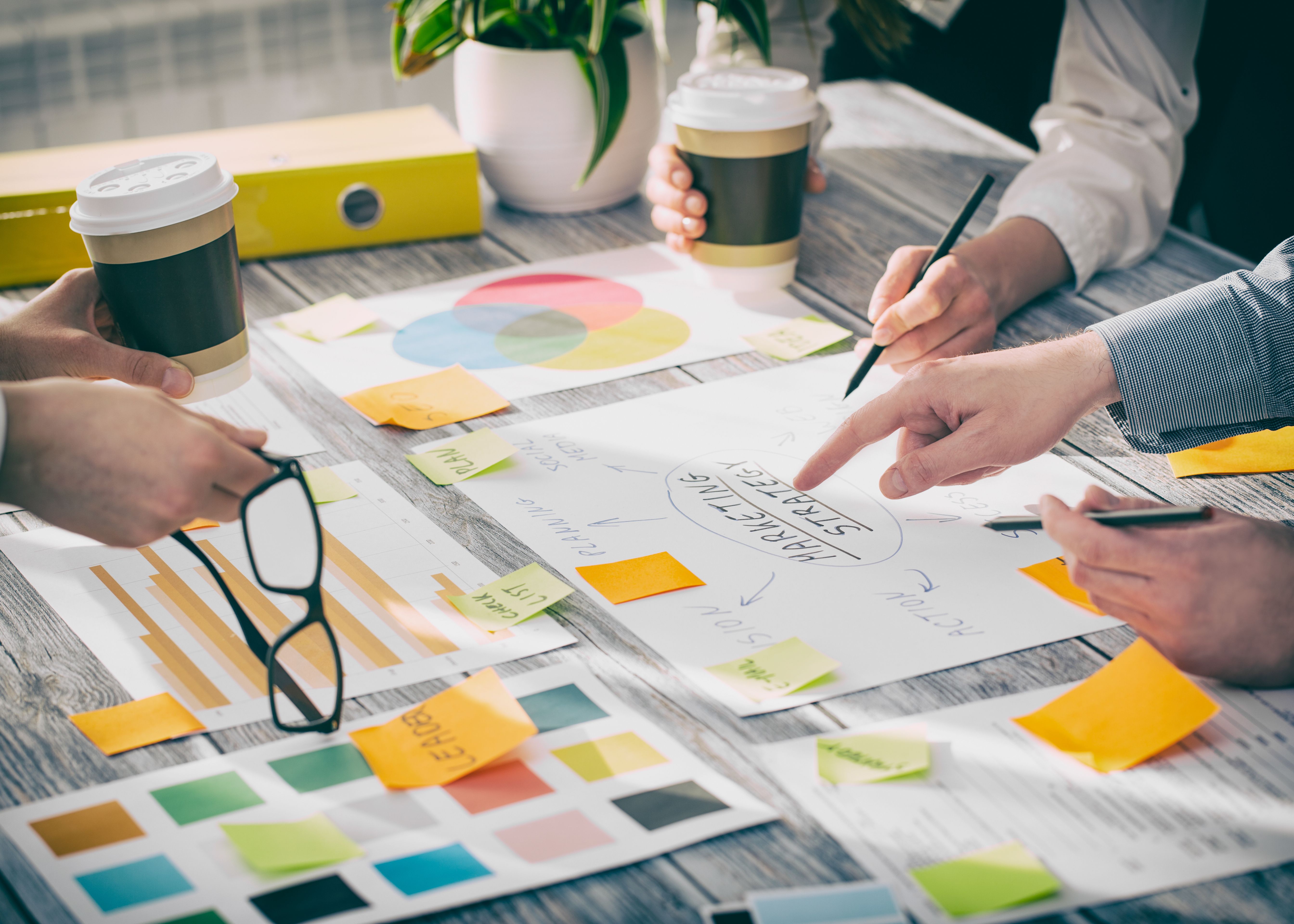 Colleagues collaborating at a desk with charts, sticky notes and takeaway coffee cups, discussing marketing strategy