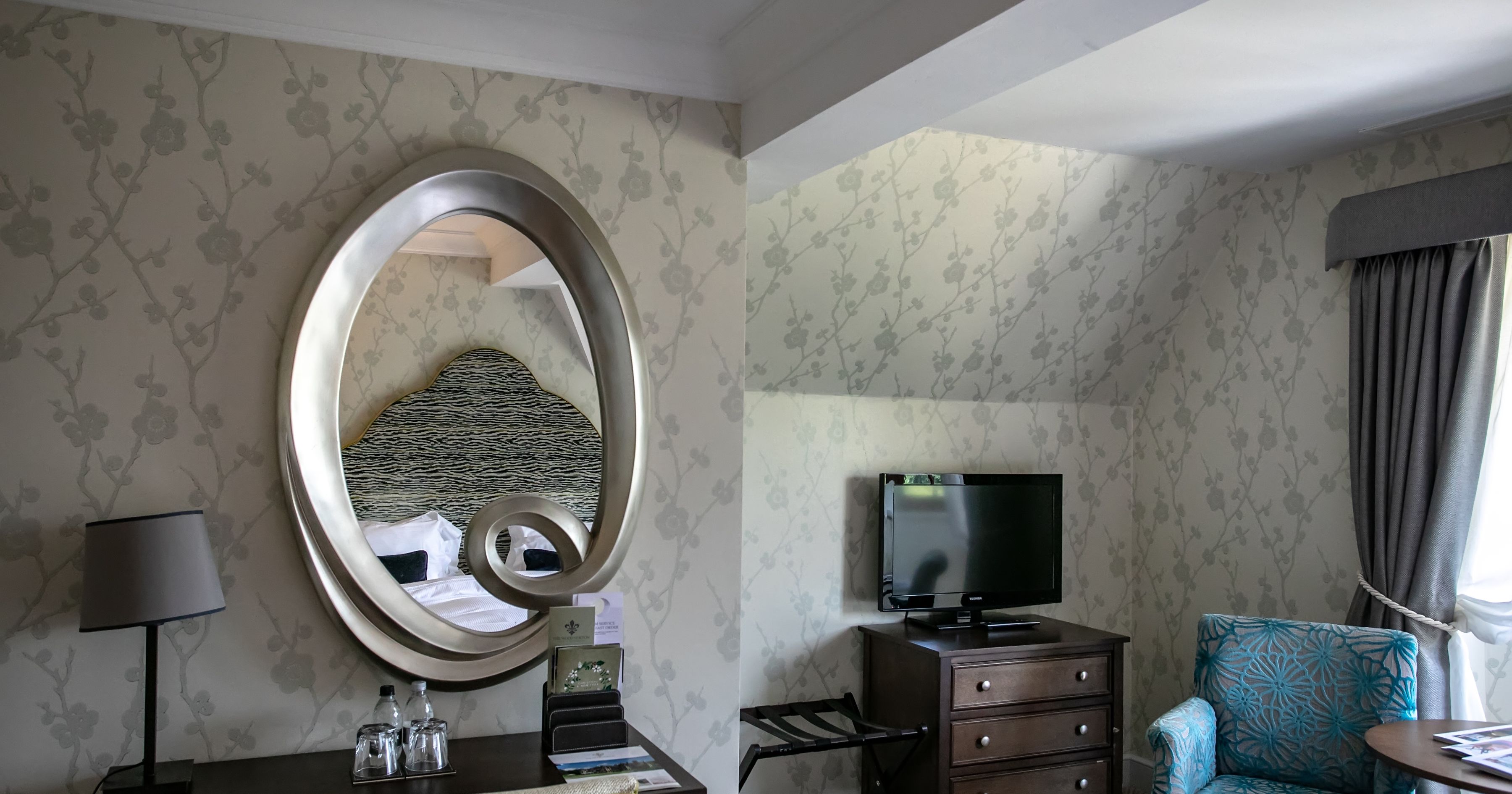 Hotel bedroom corner with ornate oval mirror above a desk, TV on a chest of drawers and an upholstered chair by the window