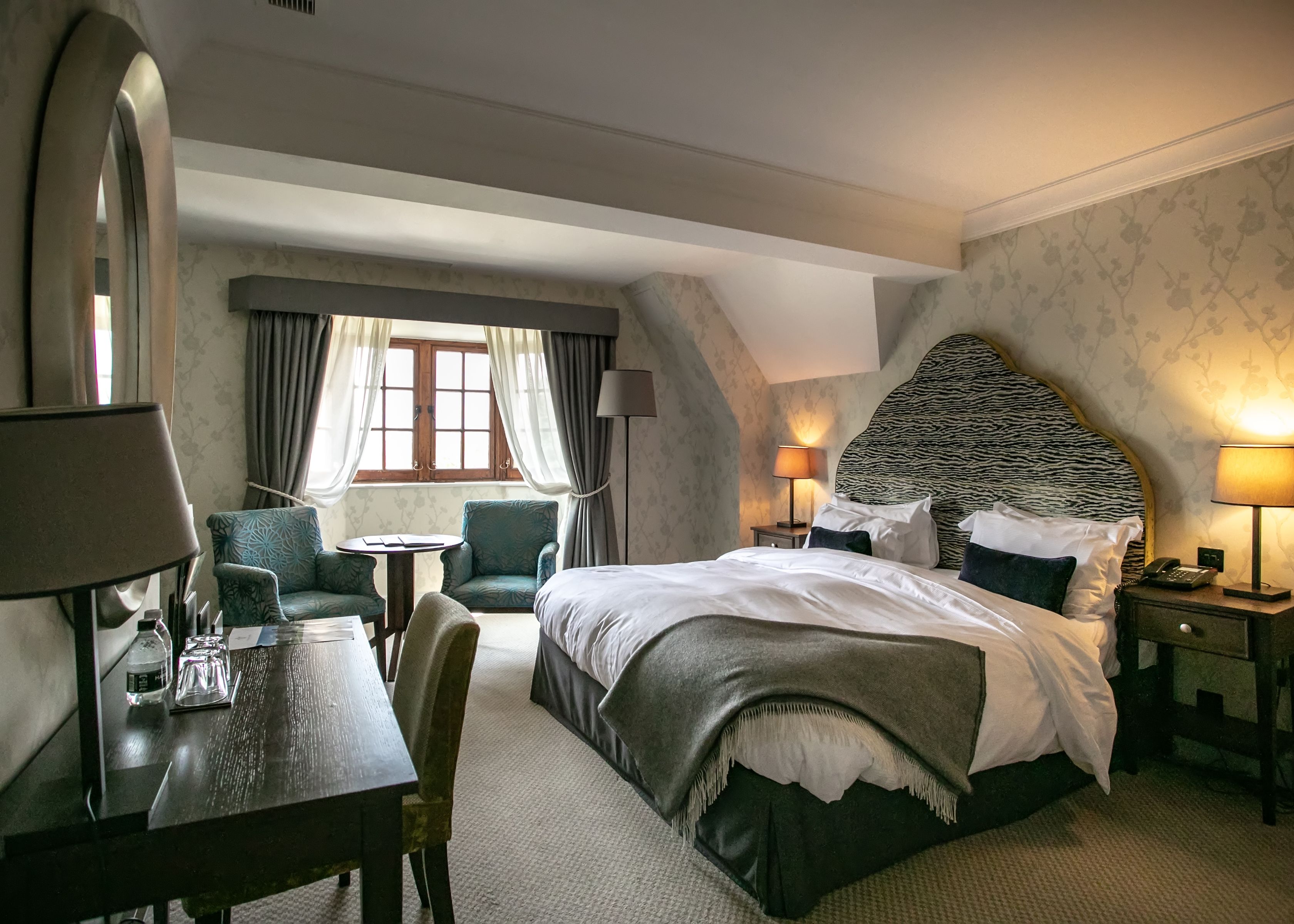 Elegant hotel bedroom with double bed, patterned headboard, bedside lamps and armchairs by a window