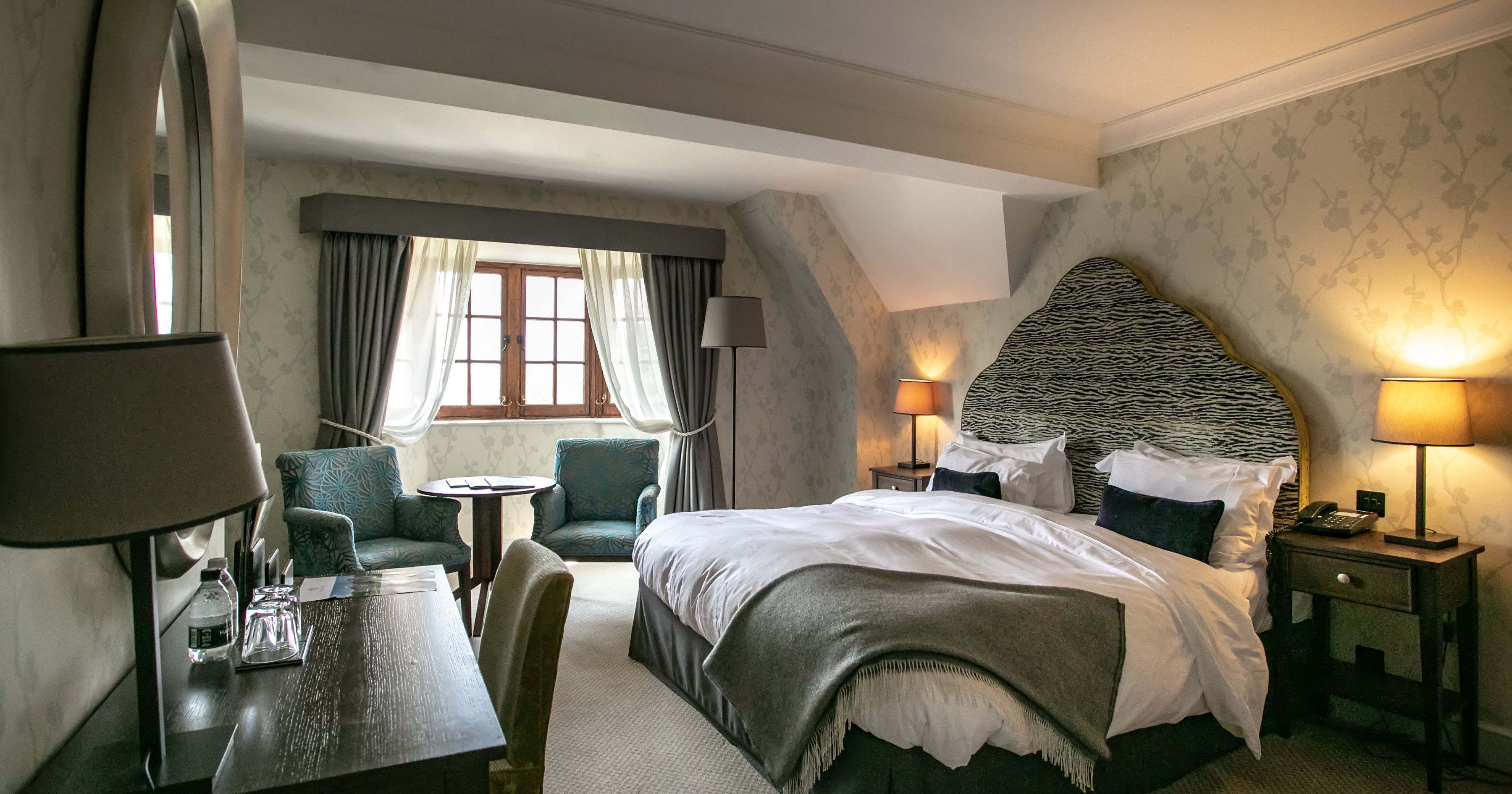Elegant hotel bedroom with double bed, patterned headboard, bedside lamps and armchairs by a window