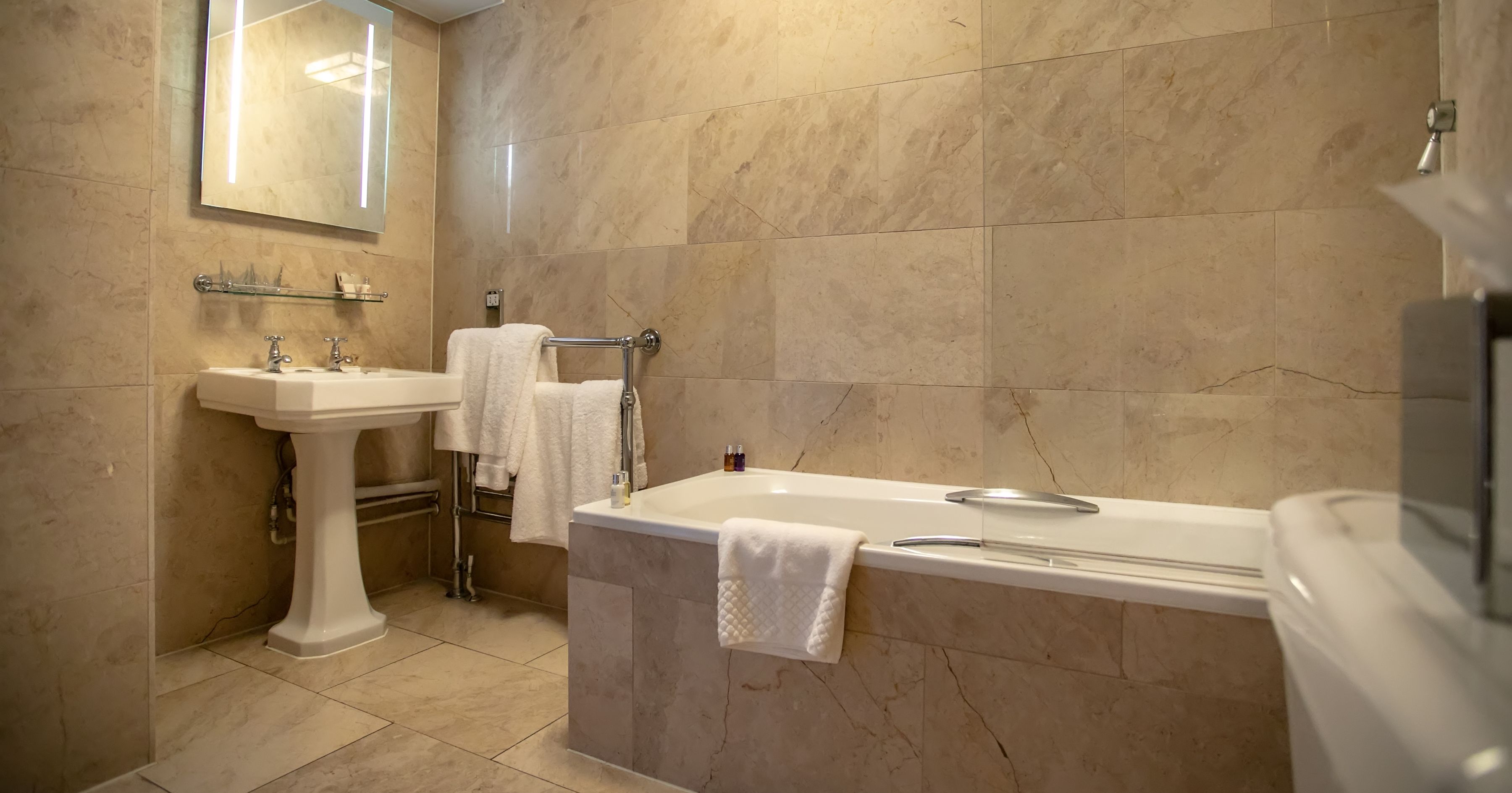 Modern hotel bathroom with marble-effect tiled walls, pedestal sink, illuminated mirror and bathtub with white towels
