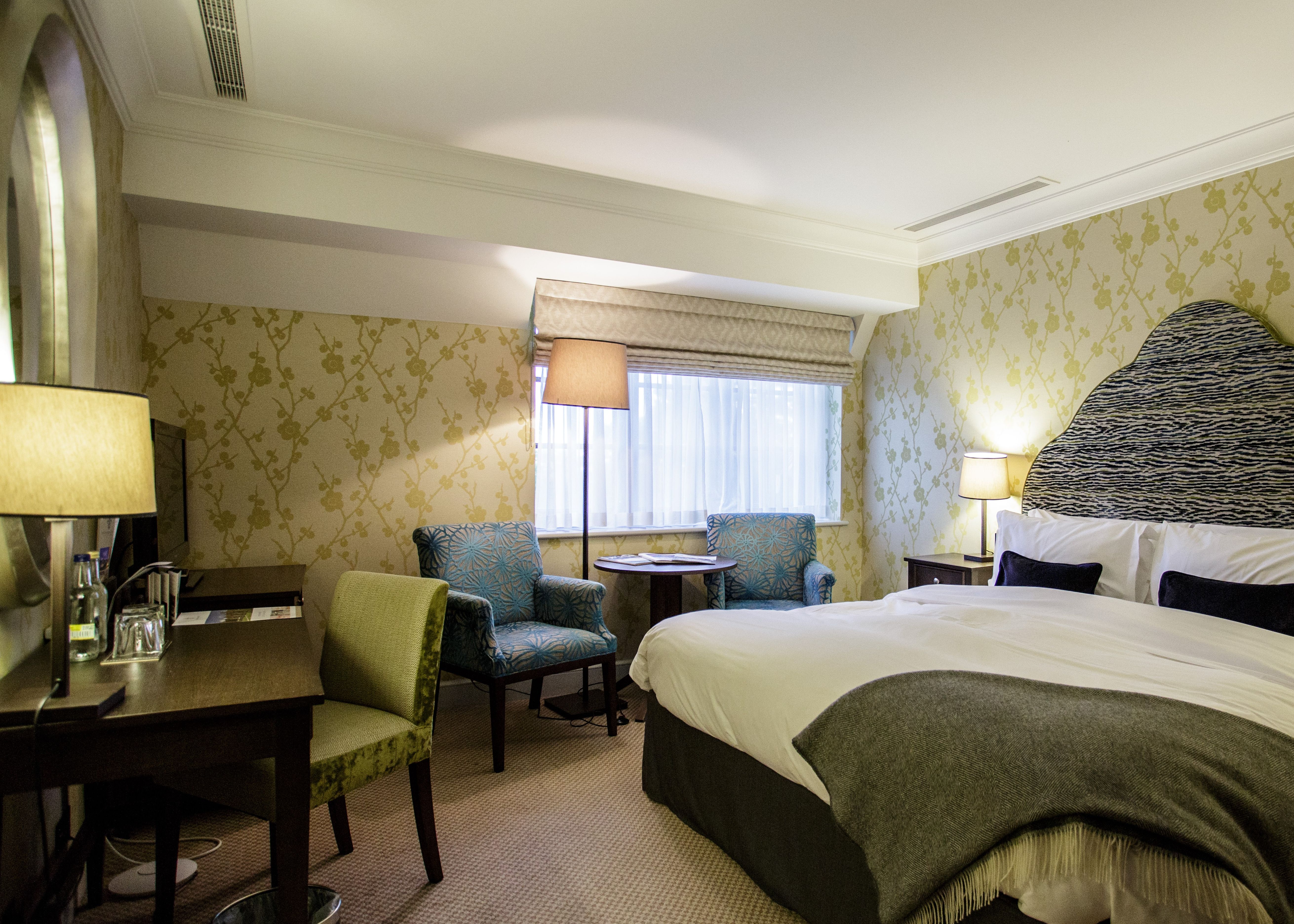 Elegant hotel bedroom with double bed, patterned headboard, bedside lamps and armchairs by a window
